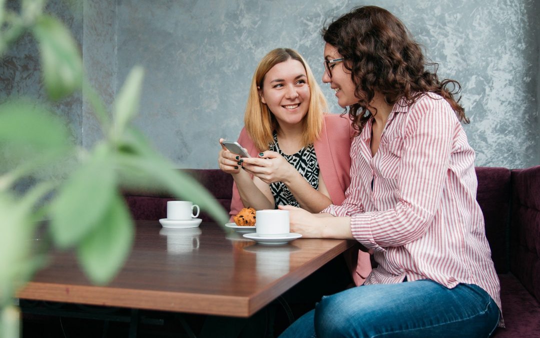 Na zdjęciu dwie młode kobiety siedzą przy stoliku w kawiarni i rozmawiają uśmiechając się do siebie. Jedna z nich trzyma telefon. Na stoliku znajdują się dwie filiżanki kawy i kawałek ciasta.