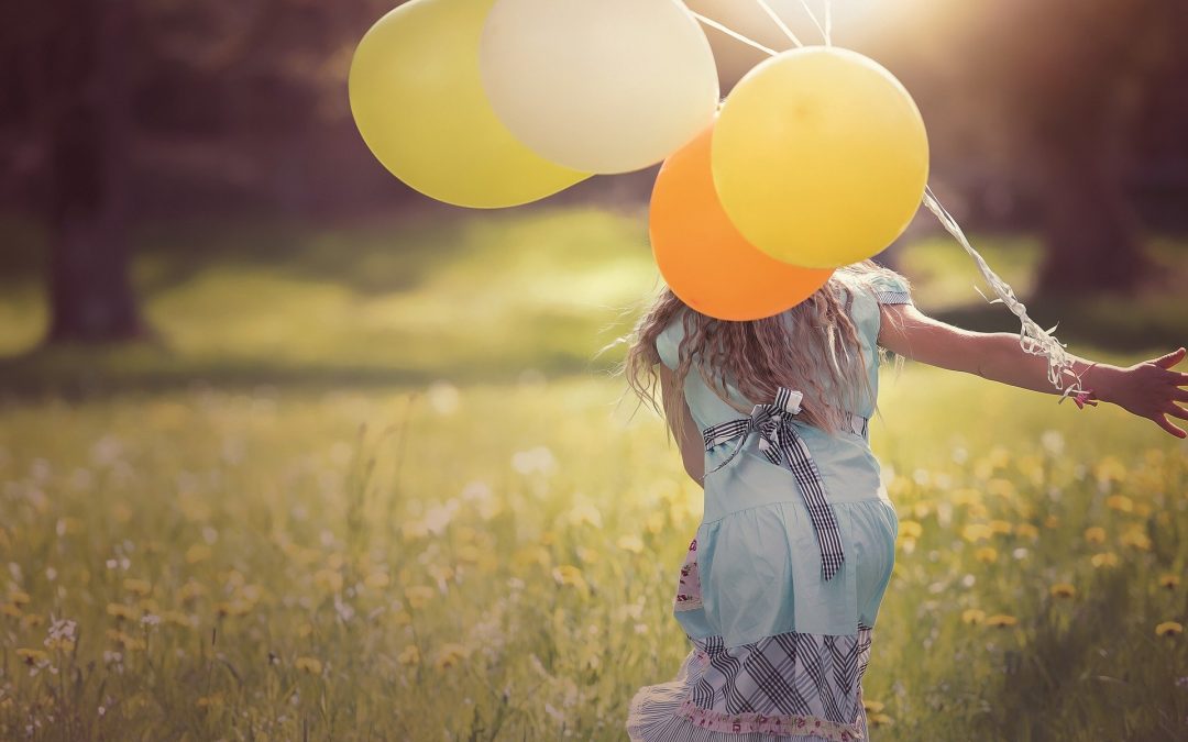 Jest słoneczny, letni dzień. Młoda dziewczyna zwrócona tyłem idzie w letniej sukience przez kwitnącą łąkę. Ma długie, blond włosy. Do prawej ręki ma przywiązany pęk Żółtych balonów.