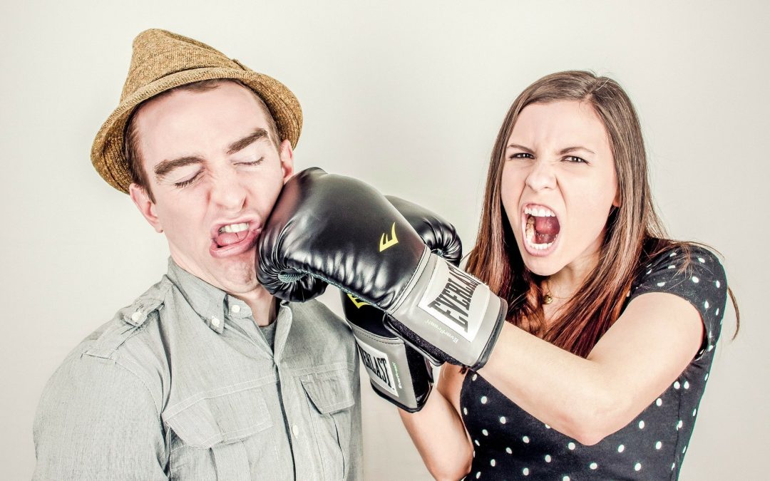 Na zdjęciu młoda kobieta i mężczyzna. Kobieta wygląda na wściekłą i uderza mężczyznę w twarz lewą ręką z założoną rękawicą bokserską. Mężczyzna się nie broni, prawdopodobnie czuje się winny z jakiegoś powodu.