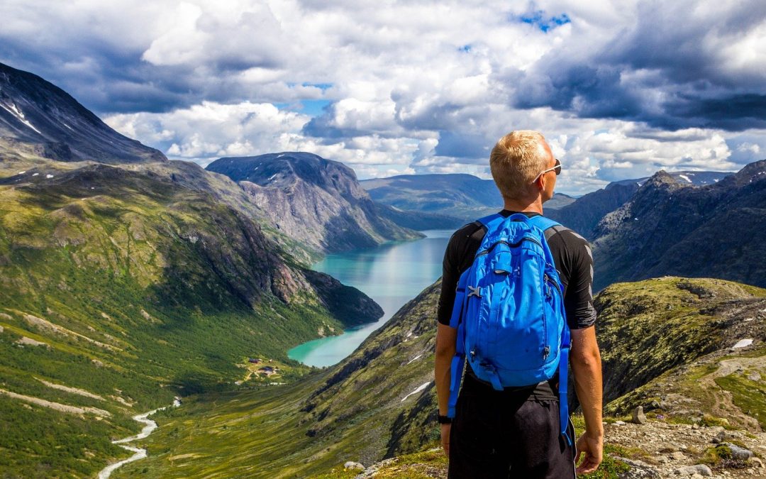 Młody mężczyzna z niebieskim plecakiem, w okularach przeciwsłonecznych stoi na wzgórzu i zastanawia się, w którą stronę pójść